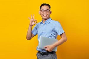 Portrait of smiling handsome Asian man in glasses holding laptop and gesturing okay sign isolated on yellow background. businessman and entrepreneur concept