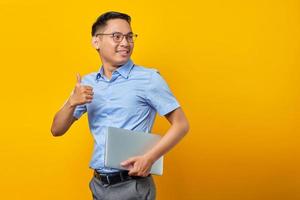 Portrait of smiling handsome Asian man in glasses holding laptop and making thumb up gesture, approve good thing isolated on yellow background. businessman and entrepreneur concept photo