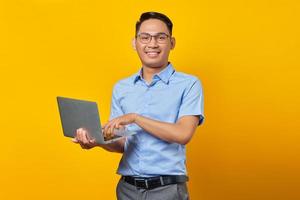 Portrait of cheerful handsome Asian man in glasses using laptop isolated on yellow background. businessman and entrepreneur concept photo