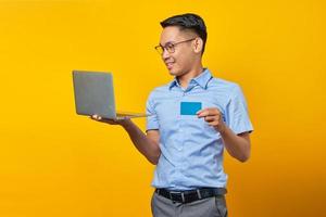 un joven asiático sonriente asiático con anteojos sosteniendo una laptop y mostrando una tarjeta de crédito aislada en un fondo amarillo. concepto de empresario y emprendedor foto