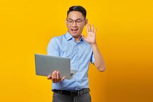 retrato de un alegre joven asiático asiático con anteojos usando una laptop, haciendo una videollamada, saludando a amigos aislados de fondo amarillo. concepto de empresario y emprendedor foto