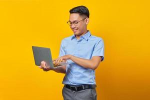 Portrait of cheerful handsome Asian man in glasses using laptop isolated on yellow background. businessman and entrepreneur concept photo