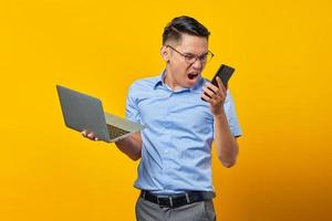 Annoyed young asian man Asian in glasses holding laptop and using a mobile phone while loudly shouting isolated on yellow background. businessman and entrepreneur concept photo