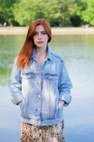 authentic young woman wearing denim jacket over summer dress standing by lake photo