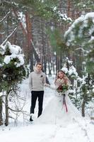 Cheerful bride and groom in beige knitted pullovers are walking in snowy forest. Winter wedding photo