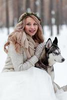 Cute bride with wreath is playing with siberian husky on background of white snow. Winter wedding. photo