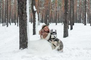 Cute bride with wreath is playing with siberian husky on background of white snow. Winter wedding. photo