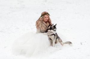 Cute bride with wreath is playing with siberian husky on background of white snow. Winter wedding. photo