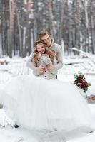 Cute couple in love with a bouquet are sitting on the log on background of the winter forest. Artwork. Winter wedding. Copy space