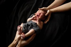 niño recién nacido sobre un fondo negro. la parte superior e inferior del niño sostienen las manos de los padres. el bebe esta llorando. foto