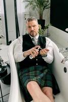 handsome mature courageous stylish man scotsman in kilt and suit laying in bathroom. Style, work from home, fashion, lifestyle, lockdown, culture, ethnic concept. photo