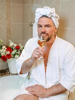 handsome mature man in white bathrobe and towel on his head sitting on bathroom with foam and drinking rose wine. Spa, relax, lifestyle, enjoy life concept.