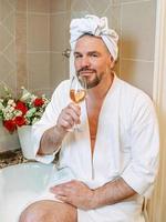 handsome mature man in white bathrobe and towel on his head sitting on bathroom with foam and drinking rose wine. Spa, relax, lifestyle, enjoy life concept. photo