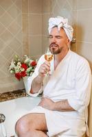 handsome mature man in white bathrobe and towel on his head sitting on bathroom with foam and drinking rose wine. Spa, relax, lifestyle, enjoy life concept.