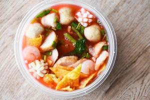 Red soup noodle pork balls crispy wontons with seafood squid shrimp and fish balls vegetable in soup bowl plastic on wooden table, top view Thai and China Asian food delivery concept photo