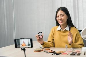 Young asian woman vlogging about cosmetics skin care items products on table with her video camera and demonstrates product use and reviews for her online blog channel. photo