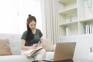 una mujer asiática se sienta en el sofá mientras toma notas de la pantalla de su computadora mientras trabaja o estudia en línea en casa. foto