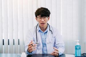médico asiático con anteojos y uniformes blancos con estetoscopio brinda asesoramiento en línea a los pacientes durante el brote de virus manteniendo una distancia social. foto