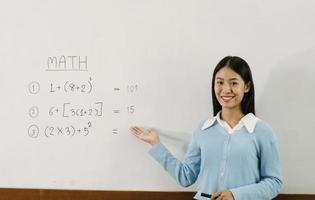 Asian female teacher is teaching students at the classroom while pointing at numbers on the white board. photo