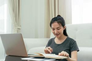 una joven asiática se sienta en la sala de estar usando una calculadora para calcular los ingresos y gastos familiares y lo escribe en su cuaderno o computadora portátil en casa. foto