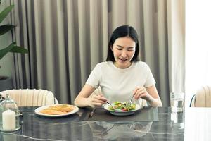 Asian young woman smiling as she scoops a salad on a plate and eats happily at home. photo