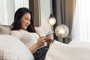 Asian woman in bed checking social apps with smartphone in morning.