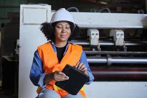 African American female engineer worker in safety uniform and hard hat, quality inspect by tablet, maintenance, check the machine at paper production manufacture industry, factory technician manager. photo