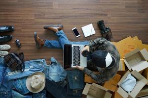 Top view of man holding coffee cup working laptop computer with fashion accessories on wooden floor from home. With postal parcel, Selling online ideas concept design photo