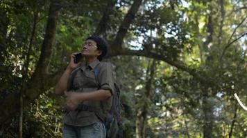 mochilero femenino usando teléfono móvil hablando con la familia en el bosque tropical. video