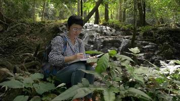 la botanica sta esaminando i dati biologici sulle piante verdi vicino alla cascata nella foresta tropicale. video