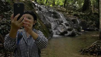 mujer excursionista en el bosque tomando una videollamada selfie en una pequeña cascada. video