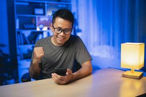 hombre asiático animando deportes mientras ve el teléfono móvil en vivo, sonriente y feliz. hombre asiático sonriendo y feliz de ganar una apuesta. animando el éxito del concepto. foto