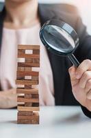 Businesswoman hand holding glass magnifying over wooden blocks tower. Business analysis, Risk Management, Study, opportunities and strategy Concepts photo