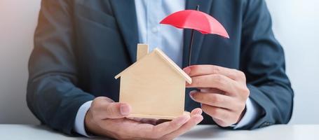 mano de hombre de negocios que sostiene el modelo de casa de madera cubierta de paraguas rojo. conceptos inmobiliarios, de seguros y de propiedad