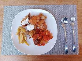 Food consisting of fried fish in tartar sauce, chili chicken, french fries, and vegetables with red oyster sauce on a white plate, with a brown wooden table mat photo