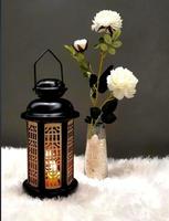 The Ramadan lantern is black, luminous, and its sides are decorated with wooden decorations, placed next to a small vase containing white crystals and carrying some white roses as well, both of which
