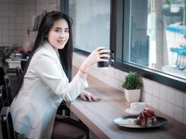 joven y bella mujer de negocios asiática sonriendo y posando mientras se sienta en una cafetería moderna. mujer joven y atractiva sentada en un café interior y mirando feliz a la cámara. foto