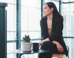 retrato de una elegante mujer de negocios asiática sentada en una cafetería con una taza de café sola mirando hacia un lado y pensando en una puesta en marcha foto