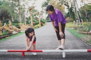 Asian mother and her daughter as they stretching their legs before they go for a run at the park. The little girl is seen from behind. Mother and daughter are wearing fitness gear.