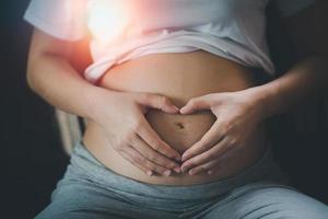 mujer embarazada cogidos de la mano en forma de corazón en el vientre en casa. mujer embarazada haciendo corazón con las manos, primer plano. foto