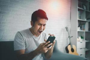 Depression concept. Asian man sad and checking mobile phone sitting on the sofa in the living room at home, Asian man serious and worry while using mobile phone. photo