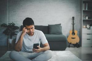 Depression concept. Asian man sad and checking mobile phone sitting on the sofa in the living room at home, Asian man serious and worry while using mobile phone. photo