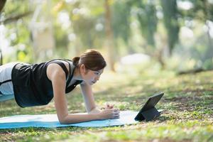 Asian fitness woman doing a yoga according to a video clip. Asian woman using tablet and doing yoga in park photo