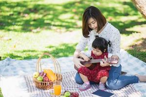 familia con niños disfrutando de un picnic en el jardín de primavera. padres e hijos divirtiéndose almorzando al aire libre en el parque de verano. madre e hija tocando el ukelele en el jardín foto