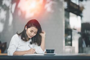 mujer asiática sentada y usando un bolígrafo para escribir en un cuaderno de papel con una computadora portátil y usando auriculares. trabajar desde cualquier lugar concepto. trabajo desde el concepto de hogar. foto