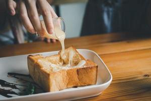 Woman pouring condensed milk on toast on the dining table, woman eating toast in the cafe restaurant. Food bakery concept. photo