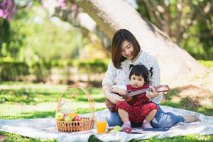 Family with children enjoying picnic in spring garden. Parents and kids having fun eating lunch outdoors in summer park. Mother and daughter playing ukulele in garden photo