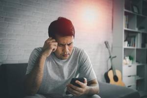 Depression concept. Asian man sad and checking mobile phone sitting on the sofa in the living room at home, Asian man serious and worry while using mobile phone. photo