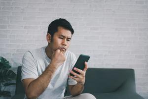 concepto de depresión. hombre asiático triste y revisando el teléfono móvil sentado en el sofá de la sala de estar en casa, hombre asiático serio y preocupado mientras usa el teléfono móvil. foto