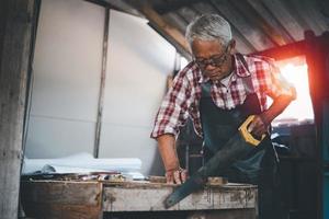 Senior old carpenter using saw working on wood craft at workshop to produce construction material or wooden furniture. photo